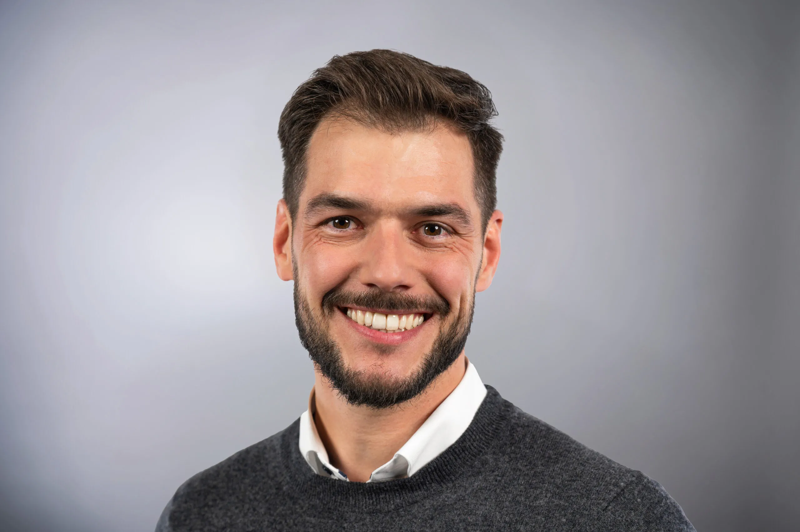 Porträt von Jens Hippchen. Er trägt einen grauen Pullover über einem weißen Hemd, der Hintergrund ist grau.