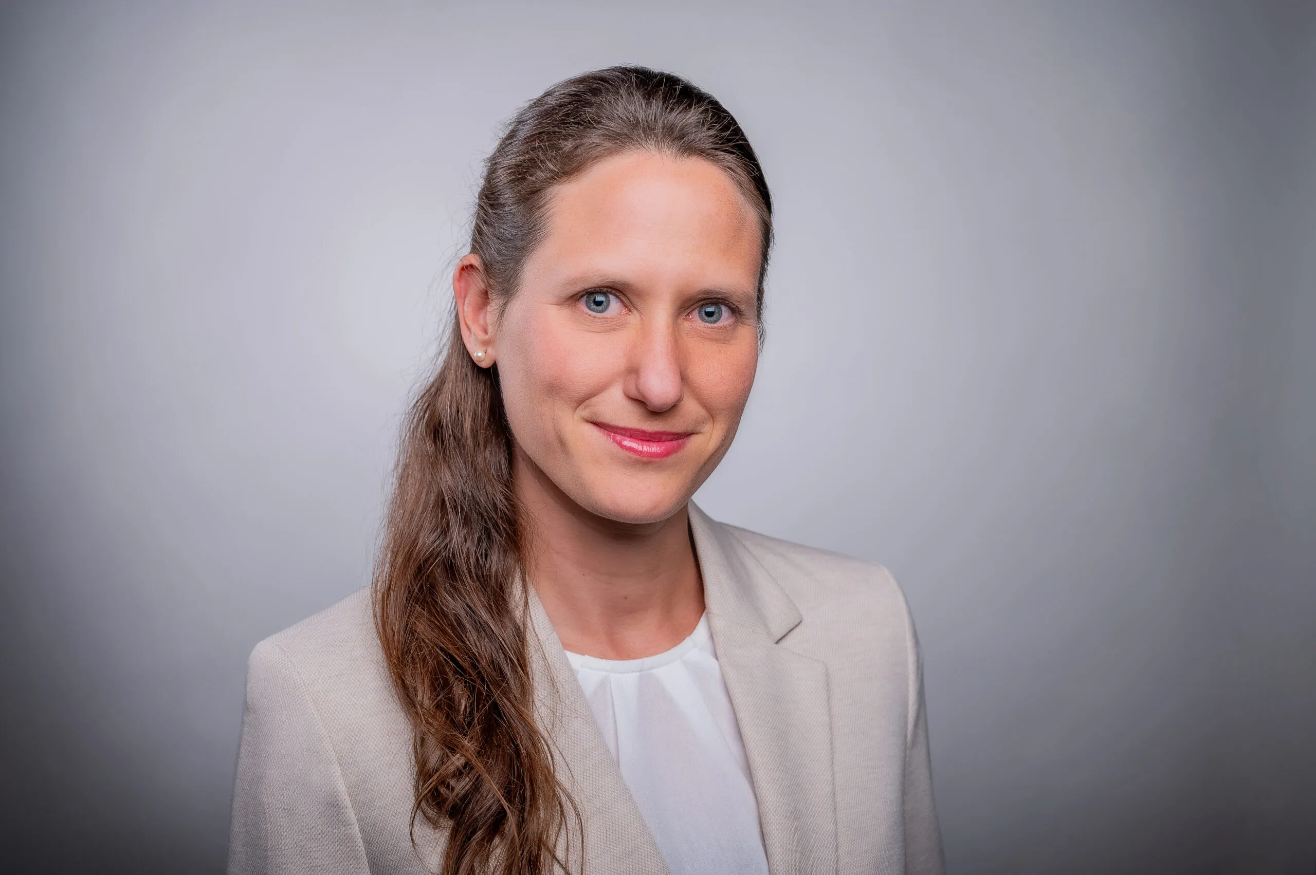 Porträt von Jasmin Völlkopf. Sie trägt die Haare zu einem Zopf gebunden, dazu einen beigen Blazer mit weißer Bluse. Der Hintergrund ist grau.